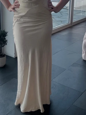 campange färgad långklänning med öppen rygg - Elegant champagne färgad långklänning med djup v-ringning och öppen rygg. Klänningen har en slät, figurnära passform och är ärmlös, vilket ger en stilren och modern look. Perfekt för speciella tillfällen när du vill sticka ut.