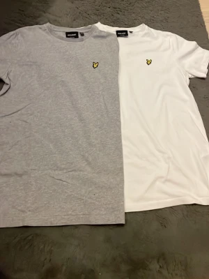 2-pack Lyle & Scott t-shirts grå/vit - Säljer två klassiska t-shirts från Lyle & Scott, en i ljusgrått och en i vitt. Båda har rund hals, korta ärmar och den ikoniska gula örnloggan broderad på bröstet. Perfekta basplagg i mjuk bomull som passar till allt.