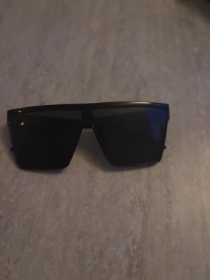 Svarta solglasögon - Säljer ett par svarta solglasögon med bred, rektangulär form och raka linjer. Glasögonen har en modern, futuristisk look med helsvart båge och mörka linser. Tillverkade i plast och perfekta för dig som vill sticka ut med en edgy stil.