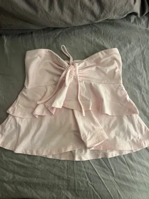 Ljusrosa bandeau topp med volang - Supersöt ljusrosa bandeau topp med volangdetaljer och knyt framtill. Toppen är ärmlös och har en mjuk, flowy passform som ger en romantisk vibe. Perfekt för sommardagar och festivaler.