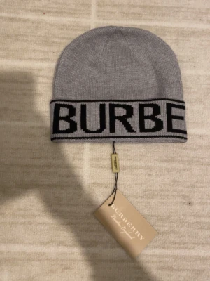 Grå mössa från Burberry med logga - Snygg grå mössa från Burberry med bred uppvikt kant och stor svart logotyp runt hela mössan. Tillverkad i mjukt stickat material som känns skönt mot huvudet. Perfekt accessoar för dig som vill ha en stilren och trendig look.