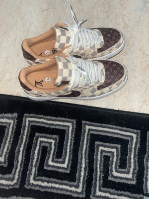 Louis Vuitton x Nike Air Force 1 lows - Unika sneakers med klassiskt rutmönster i beige och brunt samt bruna partier med Louis Vuitton-monogram. Modellen är en low-top med vita skosnören och beige innersula. Perfekt för dig som vill sticka ut med lyxig streetstyle.