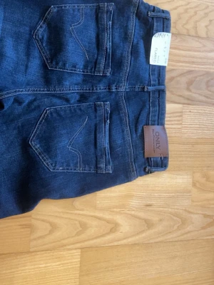 Mörkblå bootcut jeans från ONLY - Snygga mörkblå bootcut jeans från ONLY med klassiska bakfickor och bruna detaljer vid linningen. Jeansen har utsvängda ben och är tillverkade i stretchigt denimtyg som sitter skönt. Perfekta för dig som gillar en retroinspirerad look.