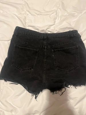 Svarta slitna jeansshorts - Säljer ett par svarta jeansshorts med råa, fransiga kanter och slitningar framtill. Shortsen har fem fickor, bälteshällor och en klassisk knappgylf. Perfekta för dig som gillar en edgy och avslappnad stil.
