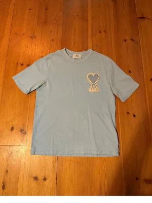 Ljusblå t-shirt från Ami Paris -  Säljer en ljusblå t-shirt från Ami med ett stort broderat hjärta och bokstaven A på bröstet. T-shirten har rund hals och korta armar, tillverkad i mjuk bomull. Perfekt för dig som gillar stilrena plagg med en twist. Den har lite gula fläckar under armhålan men det går bort.
