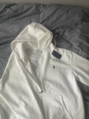 Vit hoodie från Polo Ralph Lauren - Snygg vit hoodie från Polo Ralph Lauren med dragkedja och klassisk huva. Ikoniska loggan broderad på bröstet i mörkblått. Mjuk bomullskänsla och fickor framtill. Perfekt för en clean och stilren look. Sälja pga inte passade mig