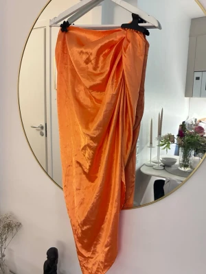 Orange omlottkjol i satin - Snygg lång omlottkjol i glansigt orange satinmaterial. Kjolen har draperad effekt baktill och en elegant, asymmetrisk form. Perfekt för dig som vill sticka ut med färg och stil.