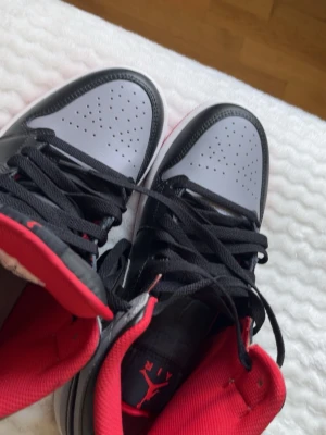 Air Jordan 1 Mid svart/grå/röd - Snygga Air Jordan 1 Mid sneakers i svart och grått läder med röda detaljer och vit sula. Klassisk Nike Swoosh på sidan och röd Jumpman-logga på plösen. Perfekta för dig som gillar streetstyle och vill sticka ut med en ikonisk modell.