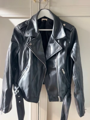 skinnjacka med bälte - Snygg svart skinnjacka i klassisk bikerstil med breda slag, silverfärgade dragkedjor och tryckknappar. Jackan har ett matchande bälte i midjan och dragkedjor vid ärmsluten. Perfekt för dig som vill ha en edgy och tidlös look. Jackan är i storlek 40 men ganska liten i modellen.