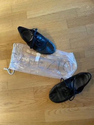 Svarta dansskor från Capezio - Svarta dansskor från Capezio i mjukt läder med snörning och låg profil. Perfekta för dans eller träning, levereras med originalpåse. Skorna har en klassisk och smidig design som ger bra rörelsefrihet. Original påsen är sliten men det finns en annan nu kan få de i om ni skulle vilja det