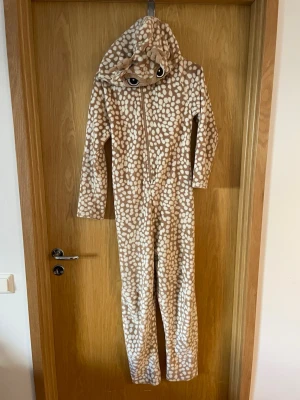 Brun prickig onesie med huva - Supermysig onesie i mjuk fleece med brunt och vitt bambi-mönster. Huvan har söta öron och broderade ögon, samt dragkedja framtill. Perfekt för chillkvällar hemma eller som rolig pyjamas.