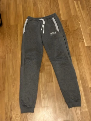Grå mjukisbyxor från Hugo Boss - Snygga grå mjukisbyxor från Hugo Boss med vita detaljer och dragsko i midjan. Byxorna har vita sidopaneler, ribbade muddar vid benslut och logga tryckt på vänster lår. Perfekta för en chill och avslappnad stil.