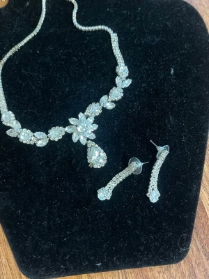 Glittrande smyckeset med strass - Säljer ett elegant smyckeset med halsband och matchande örhängen, dekorerade med gnistrande klara strass-stenar i blommönster och droppform. Perfekt för dig som vill ha extra bling till din outfit. Silverfärgad metall och klassisk design.