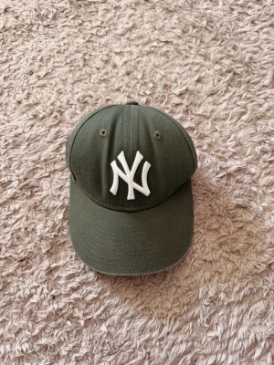 Olivgrön New Era NY keps (barnstorlek) - Snygg olivgrön keps från New Era med klassisk böjd skärm och broderad vit NY-logga framtill. Modellen är 9FORTY och har justerbar rem bak för perfekt passform. Tillverkad i slitstark bomull, perfekt för en sportig och trendig look.