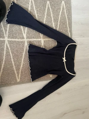 Mörkblå ribbad topp med rosett - Säljer en mörkblå croppad topp med långa ärmar och vit kant runt halsringningen. Toppen har en söt liten rosett framtill och vågiga kanter vid ärmslut och nederkant. Ribbad struktur för en trendig look.