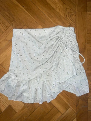 Vit blommig kjol med volang - Supersöt vit kortkjol med små blommor, volangkant och snörning i sidan för en snygg omlotteffekt. Perfekt för varma dagar och ger en romantisk vibe till din outfit.
