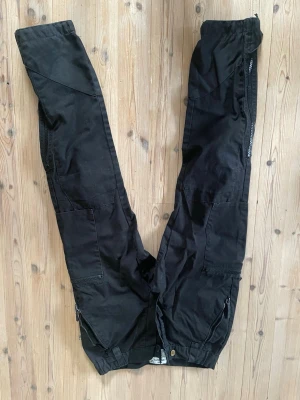 M90 militärbyxa - Svarta cargobyxor från Fjällräven med flera praktiska fickor och dragkedjor. Byxorna har en avslappnad passform och är tillverkade i slitstarkt material. Perfekta för dig som gillar outdoor och streetstyle. 