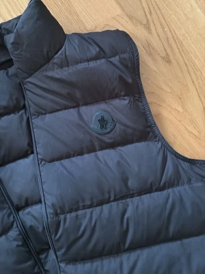 Svart dunväst från Moncler - Snygg svart dunväst från Moncler med diskret logotyp på bröstet. Västen har ståkrage, två dragkedjeförsedda fickor och quiltad design. Perfekt för lager-på-lager och håller dig varm med stil. Materialet är glansigt och känns lyxigt. Storlek 2/M
