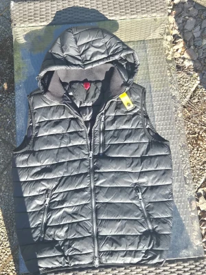 Svart parajumper dunväst med huva och dragkedjor - Snygg svart dunväst från para jumper med huva och två dragkedjefickor framtill. Västen har en gul detalj vid axeln och quiltad design. streetstyle. Materialet är glansigt och lätt vadderat för extra värme. Ovanlig väst varit inne på kemtvätt innan försäljning. Och blivit värderad till 1200-2000kr