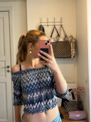 Blå offshoulder topp från KappAhl - Snygg offshoulder topp från KappAhl i blått och vitt med zickzack-mönster. Toppen har trekvartsärm och elastisk kant nertill och upptill för en skön passform. Perfekt för dig som gillar trendiga och bekväma plagg.