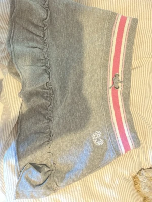 Grå shorts med rosa ränder och volang - Grå mjuka shorts från Girls League med bred resår i midjan, rosa och vita ränder samt snörning. Volangdetalj längs benen och broderad logga framtill. Perfekta för en sportig och avslappnad stil.