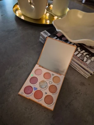 Ögonskuggspalett med spegel - Palett med nio ögonskuggor i olika nyanser av rosa, lila, guld, brunt och glitter. Paletten har inbyggd spegel och hjärtformade detaljer runt skuggorna. Färgerna är både matta och skimrande. Perfekt för kreativa sminkningar. Från colourpop. Testad , använd engång. 