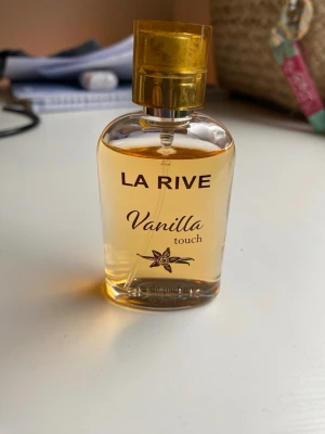 La Rive Vanilla Touch parfym - La Rive Vanilla Touch är en eau de parfum med söt vaniljdoft. Flaskan är genomskinlig med guldfärgat lock och innehållet har en ljusgul ton. Förpackningen är ljus med guldiga detaljer och en vaniljblomma på framsidan. Perfekt för dig som gillar varma, söta dofter.