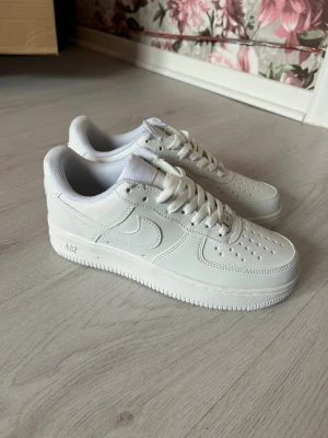 Nike Air Force 1 vita sneakers - Klassiska helvita Nike Air Force 1 sneakers i lågt utförande. Skorna har en stilren design med perforerad tå, vita skosnören och den ikoniska swoosh-loggan på sidan. Tillverkade i läder med en rejäl gummisula för skön komfort och streetig look.