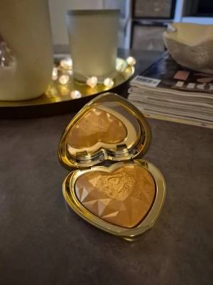 Too Faced hjärtformad highlighter - Highlighter från Too Faced i en hjärtformad guldförpackning. Pudret har en skimrande guldig nyans och en facettslipad yta som ger extra glow till din makeup. Perfekt för att framhäva ansiktets höga punkter och skapa en strålande look. Färg You light up my life. Testad, goldenglow! Lyxig förpackning!