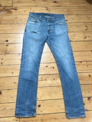 Blå raka jeans från Nudie Jeans - Snygga blå jeans från Nudie Jeans med raka ben och klassisk femficksdesign. Jeansen har slitningar och en liten reva på vänster lår samt Nudie Jeans signaturbrodyr på bakfickorna. Tillverkade i denim med en cool tvättad look. Modellen är grim Tim. Pris kan diskuteras