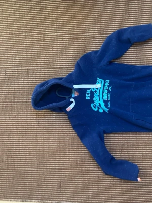 Blå Superdry hoodie med tryck - Snygg blå hoodie från Superdry med turkost tryck och japanska tecken på bröstet. Klassisk modell med huva och dragsnören, samt mjukt material som känns skönt mot huden. Perfekt för en avslappnad streetwear-look.