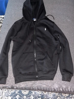 Svart zip hoodie från Polo Ralph Lauren - Svart hoodie med dragkedja från Polo Ralph Lauren. Klassisk design med huva och dragsko, två stora fickor framtill och vit broderad logga på bröstet. Mjuk bomullsblandning och ribbade muddar vid ärmslut och midja.