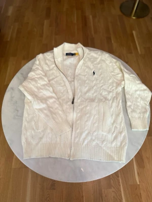 Vit kabelstickad kofta från Polo Ralph Lauren - Kabelstickad vit kofta från Polo Ralph Lauren med dragkedja framtill och ribbade muddar. Klassisk design med två fickor och den ikoniska mörkblå logotypen broderad på bröstet. Perfekt för lager på lager och en clean look.