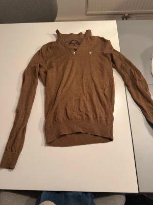Brun stickad tröja med dragkedja från Morris - Snygg brun stickad tröja från Morris med liten dragkedja vid halsen och broderad logga på bröstet. Tröjan har långa ärmar, ribbade muddar och är tillverkad i ett mjukt, tunt material. Perfekt för lager-på-lager och stilrena outfits.