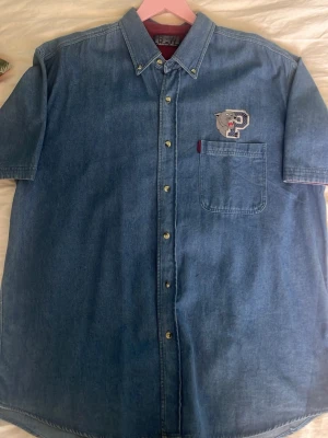 Vintage Denimskjorta  - Vintage denimskjorta med broderad panther-logo på bröstet. Klassisk 90s passform med en skön oversized känsla med snygga detaljer. Svin nice att ha över en tröja eller som stjärnan av outfiten med ett par sköna baggy jeans. Skriv gärna för mer bilder!