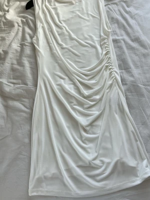 Vit klänning  - Säljer en vit midiklänning med snygga draperingar längs sidan. Klänningen är ärmlös och har en mjuk, stretchig känsla i tyget som ger en smickrande passform. Perfekt för dig som gillar minimalistisk och stilren design.