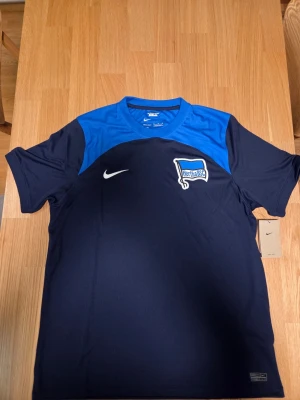 Nike Hertha Berlin fotbollströja, säsong 22/23 storlek XL - Fotbollströja från Hertha BSC. Tröjan har en mörkblå bas med blå detaljer över axlarna och övre delen av ryggen. Hertha BSC-logotypen är placerad på bröstet, tillsammans med en Nike-logotyp. Tröjan har korta ärmar och en rund halsringning.