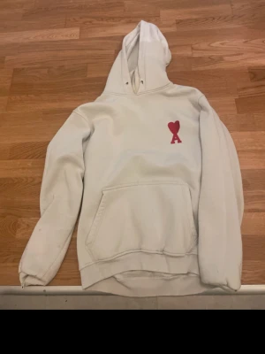 Vit hoodie från Ami Paris med rött hjärta - Vit hoodie från Ami Paris med klassisk känguruficka och huva. På bröstet sitter Ami Paris ikoniska röda hjärta med bokstaven A. Hoodien har ribbade muddar och är gjord i mjuk bomull för maximal komfort. Perfekt för en avslappnad och trendig look.