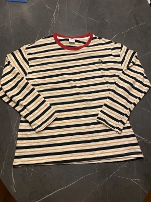 Randig långärmad tröja M – broderad orb-logga – röd krage (unisex) - Säljer en långärmad tröja från Vivienne Westwood med svarta, beige och vita ränder samt röd krage. Tröjan har broderad logga på bröstet och är tillverkad i mjukt bomullsmaterial. Perfekt för dig som gillar streetstyle och unika detaljer.