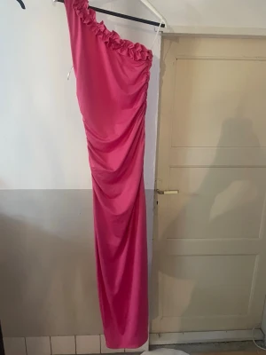 Rosa axelbandslös maxiklänning - Säljer en lång, rosa maxiklänning med en axel och volangdetalj längs urringningen. Klänningen har draperad effekt längs sidan och är i ett glansigt material som ger en elegant look. Perfekt för speciella tillfällen.