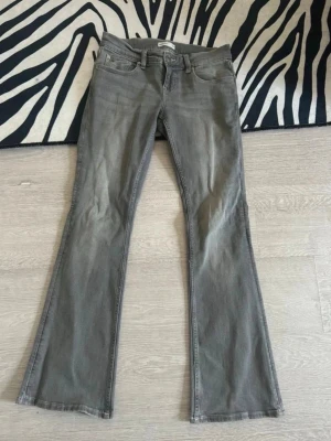 Grå bootcut jeans med låg midja - Säljer ett par grå bootcut jeans med låg midja och klassisk femficksdesign. Jeansen har en lätt utsvängd passform nedtill och är tillverkade i stretchigt denimtyg för skön känsla. Perfekta för dig som gillar retrovibbar och en avslappnad look. Från gina tricot i storlek 38. 