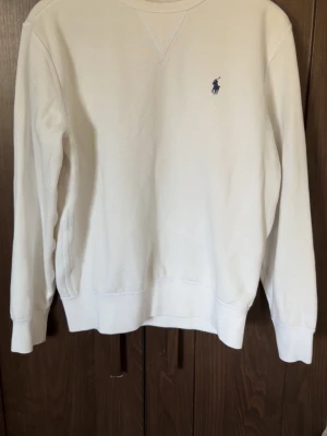  Ralph Lauren sweatshirt - Klassisk vit sweatshirt från Polo Ralph Lauren med rund halsringning och diskret blå logga broderad på bröstet. Tröjan har ribbade muddar vid ärmslut och nederkant. Perfekt för en clean och stilren look.
