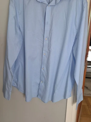 Ljusblå skjorta från H&M, storlek L - Klassisk ljusblå skjorta från H&M i storlek L. Skjortan har lång ärm, knappar framtill och en stilren krage. Perfekt för en clean och enkel look. Tillverkad i mjuk bomull som känns skön mot huden.