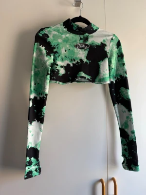 Grön och svart croppad långärmad topp - Säljer en ribbad croppad topp med grön, svart och vit tie-dye effekt. Toppen har lång ärm med tumhål och hög hals. Perfekt för dig som gillar en edgy och sportig stil. Endast provad, katt finns i hem!
