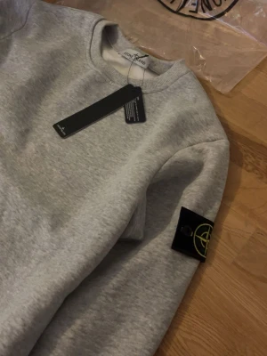 Grå sweatshirt från Stone Island - Snygg grå sweatshirt från Stone Island med klassisk rund hals och ribbade muddar. Tröjan har den ikoniska Stone Island-loggan på vänster ärm. Tillverkad i mjukt bomullsmaterial som är skönt mot huden. Perfekt för en avslappnad och trendig stil.