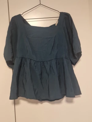 Mörkgrön blus med puffärmar och knyt - Snygg Mörkgrön blus med puffiga trekvartsärmar och knyt i ryggen. Blusen har en lös passform och en lätt glansig yta som ger en lyxig känsla. Perfekt att styla med jeans eller kjol för en trendig look.
