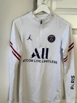 Vit PSG träningströja med dragkedja - Snygg vit långärmad träningströja från PSG och Jordan med halv dragkedja framtill. Tröjan har röda och blå detaljer på ärmarna, PSG-logga på bröstet och texten 'Accor Live Limitless' samt 'Visit Rwanda' på ryggen. Perfekt för fotbollsträning eller streetwear.