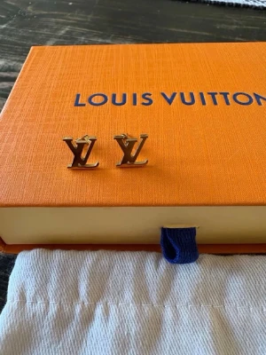 Louis Vuitton guldiga LV örhängen - Säljer ett par stilrena örhängen från Louis Vuitton i guldfärgad metall. Örhängena har den ikoniska LV-logotypen som form och kommer med original ask och tygpåse. Perfekta för dig som gillar lyxiga accessoarer och vill sticka ut med en klassisk designerstil.