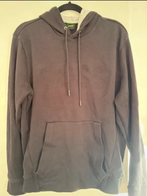 Mörkgrå hoodie från BOSS - Snygg mörkgrå hoodie från BOSS med broderad logga på bröstet. Hoodien har en stor känguruficka framtill, dragsko i huvan och mjukt grått innerfoder. Perfekt för en avslappnad och trendig stil.
