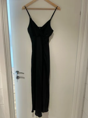 Svart långklänning från Zara - Svart långklänning från Zara med smala axelband och omlott upptill. Klänningen har en elegant v-ringning och en rak, lång siluett som ger en stilren look. Perfekt för dig som gillar minimalistisk och tidlös stil.